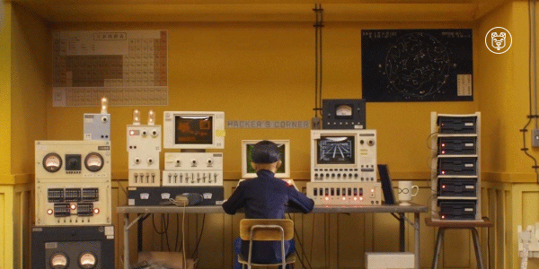 Trecho de animação que mostra um rapaz japonês sentado de costas para a câmera. Em sua frente, há uma escrivaninha com muitos equipamentos e computadores. Ele olha para a câmera, que dá um zoom, focando no rosto dele, com a tela do computador mostrando código escrito em verde sobre fundo preto. O recorte é do filme Ilha de Cachorros.