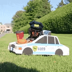 Vídeo que mostra um cachorro preto, da raça Daschund, com um quepe de policial e 'vestindo' um carro de polícia de papelão. Ele está parado em um canteiro gramado e, ao ver uma van escrito 'dogpost' na via,  sai correndo atrás.