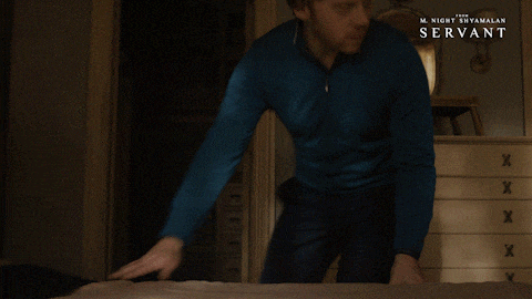 Cena do filme 'Servant' que mostra um homem ruivo vestindo camisa azul de mangas longas ajoelhando ao lado de uma cama e puxando uma maleta que estava escondida.