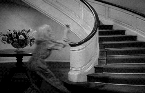 Cena de filme antigo, em preto e branco, em que uma mulher vestindo pijamas listrados sobe uma longa escada circular deslizando corrimão acima.