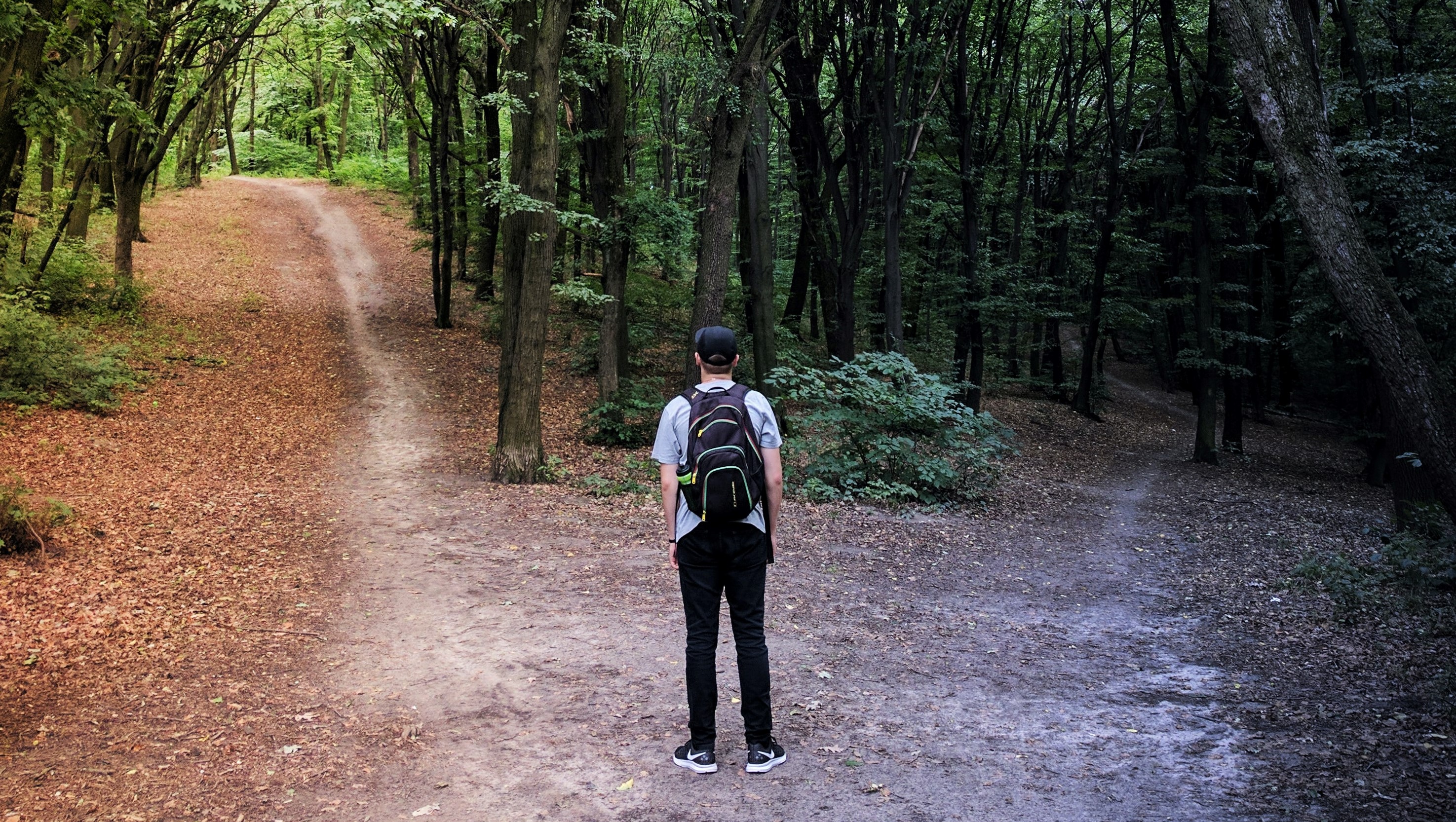 Foto que mostra um rapaz de costas, vestindo calça, camiseta, tênis, boné e mochila. À sua frente estão duas trilhas de terra  em meio às árvores e que vão para direções diferentes.
