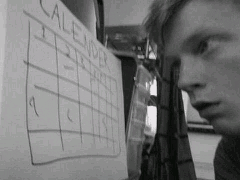 Imagem animada em preto e branco mostrando um rapaz branco, com os olhos atentos, rabiscando um calendário com uma caneta.